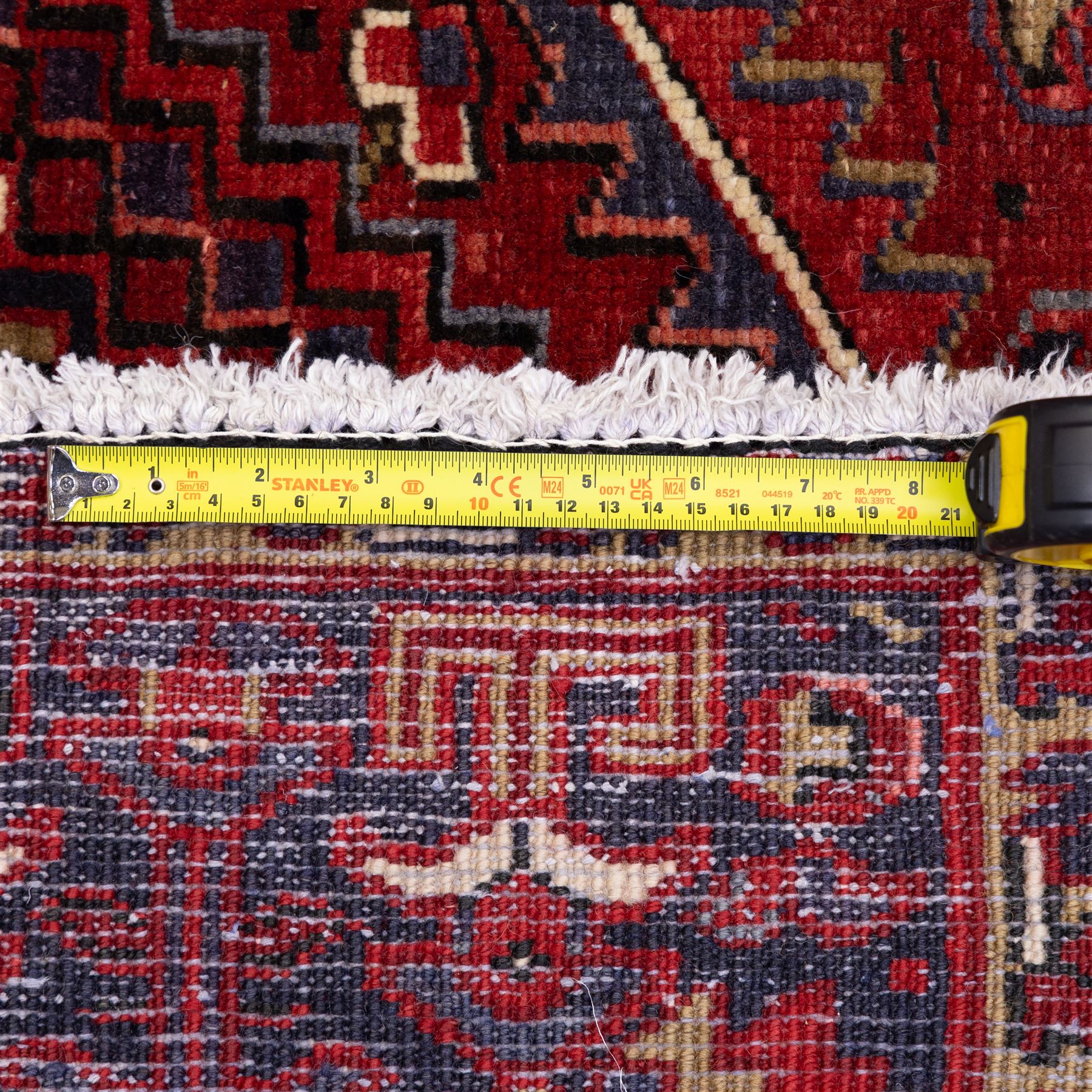 North West Persian Heriz hand-knotted carpet, madder red field with central bold angular steeped medallion with serrated outlines, flanked by stylized palmettes, hooked leaves and geometric motifs, spandrels similarly decorated within a wide geometric border and multiple guard borders in tones of madder red, indigo blue, ivory and olive green, with cotton fringe ends  