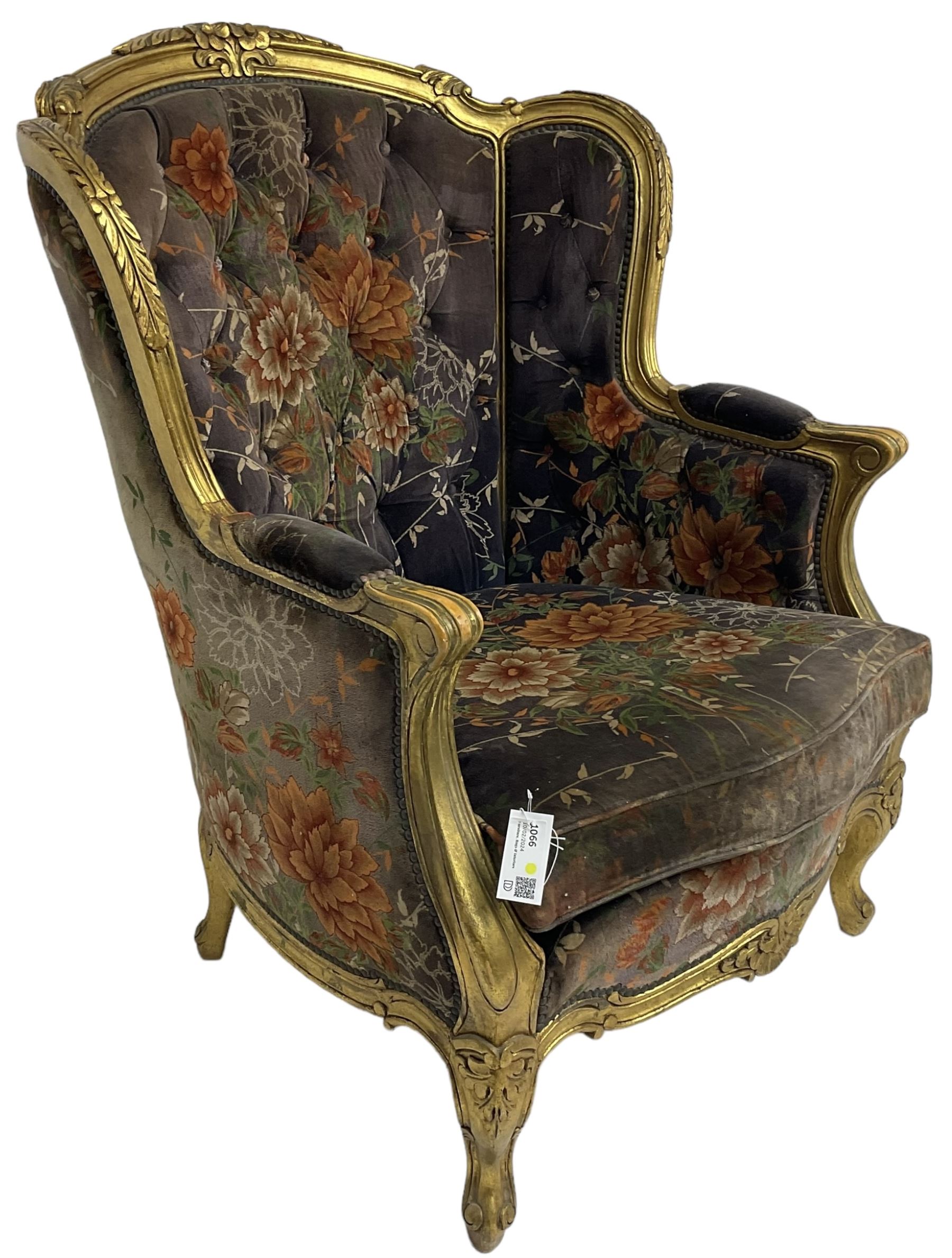 Pair of Louis XVI design gilt framed wingback armchairs, cresting rail carved and moulded with foliate decoration and C-scrolls, upholstered in buttoned floral patterned mauve velvet with loose seat cushion, scallop carved apron over cabriole supports