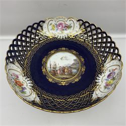 Meissen style pedestal dish, with central panel depicting a trading scene in a harbour setting, the bowl with openwork lattice sides divided by three hand painted floral panels, upon a blue ground with gilt detailing, upon a fluted knopped pedestal and circular foot, H22cm