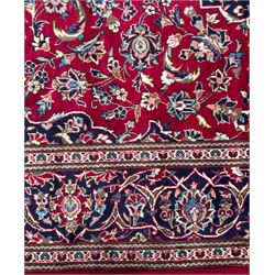 Central Persian Kashan hand-knotted carpet, the deep red ground with a central lozenge medallion in ivory and navy, surrounded by dense scrolling floral and vine motifs, navy spandrels and a wide border with stylised palmettes and flowerheads enclosed by multiple guard stripes