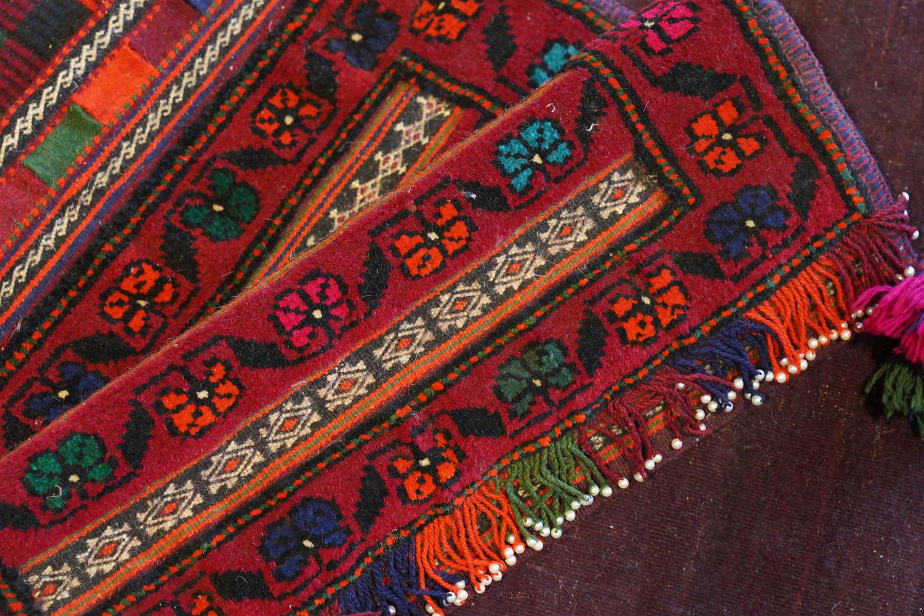 North East Persian Turkoman saddle bag, traditional double pouch form, each panel with madder red ground, decorated with horizontal bands of stylized geometric floral motifs and angular vine work, framed simple borders, central panel woven in flatweave with polychromed chequered design flanked by braided carrying cords with tasselled and knotted decorative elements 