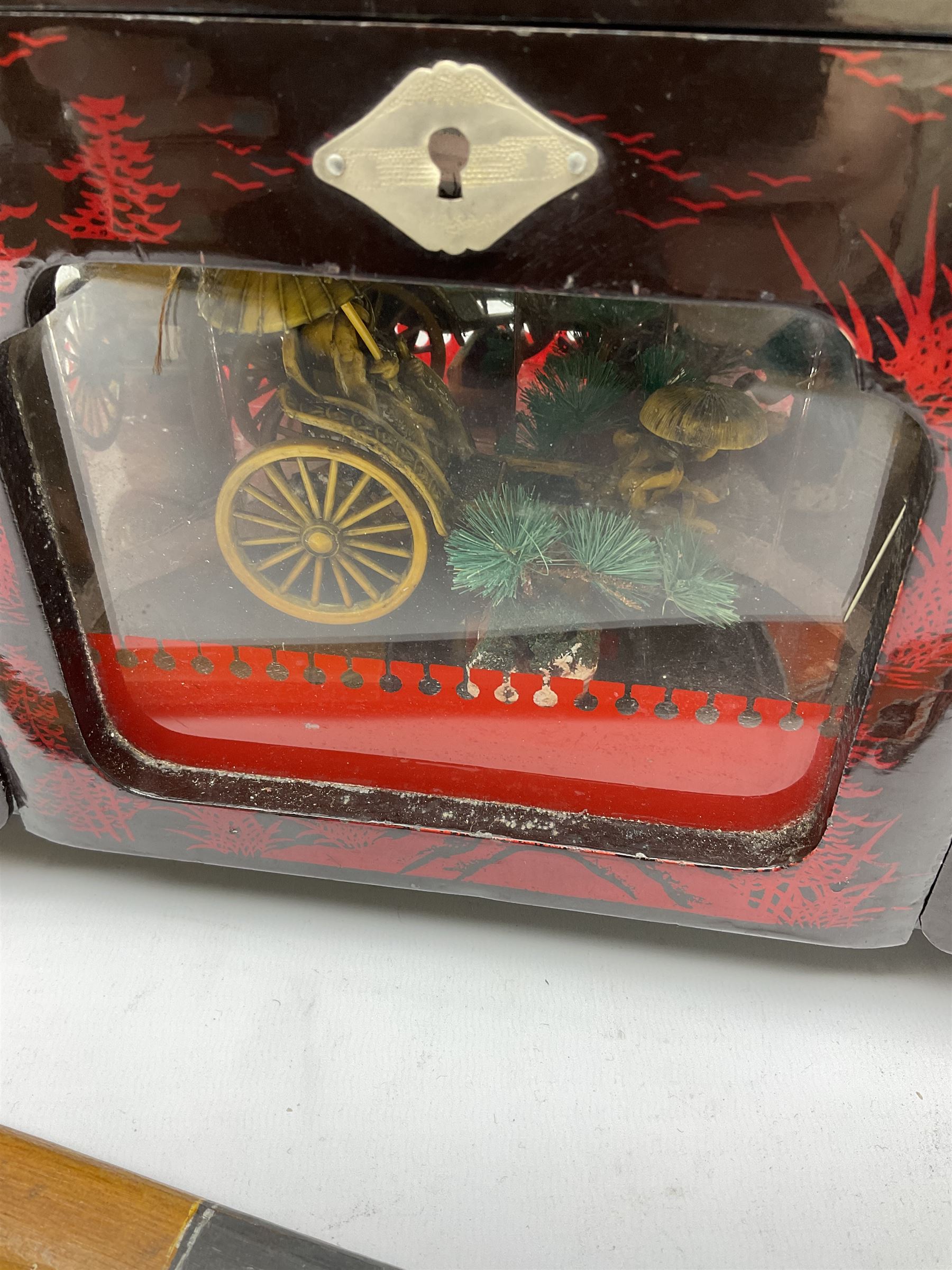 Oriental lacquered jewellery box, the body decorated with a black and red mountainous riverscape enclosing a glass compartment with simulated ivory rickshaw figure and three hinged lids opening to reveal mirror and various compartments and painted ceremonial stick