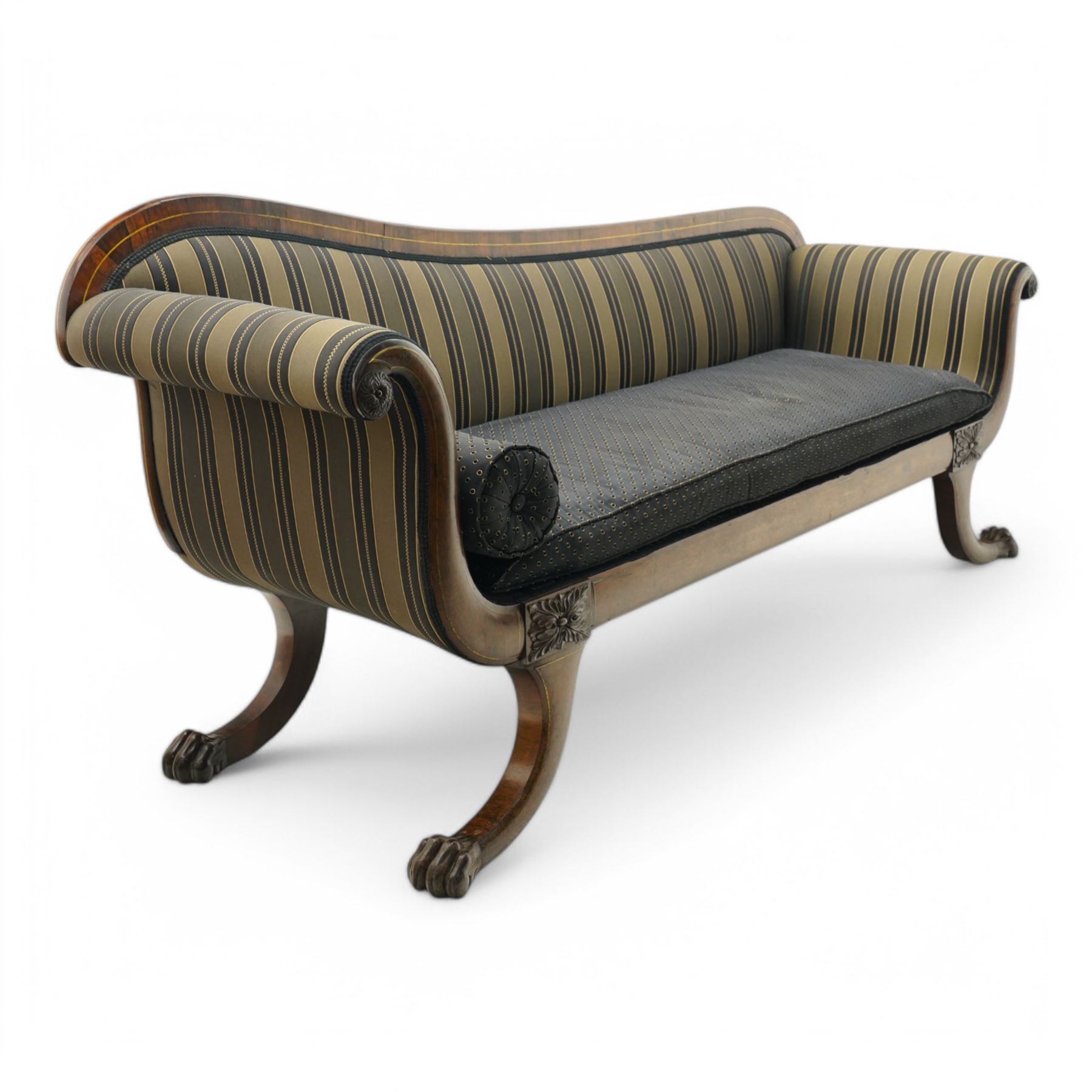 Regency brass inlaid rosewood chaise lounge or settee, shaped back over rolled head and foot, upholstered in black and gold striped fabric with rectangular squab cushion and bolster cushion, the rolled terminals carved with acanthus leaf roundels, lower rectangular foliate carved panels, on out splayed supports with paw carved feet 