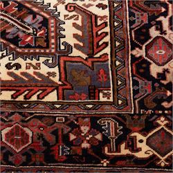 North West Persian Heriz hand-knotted carpet, madder red field with central stepped geometric medallion with radiating angular motifs and serrated outlines, and stylized floral and tribal devices framed by a broad border of repeating angular palmettes and rosettes flanked by multiple guard stripes, with cotton fringe ends 
