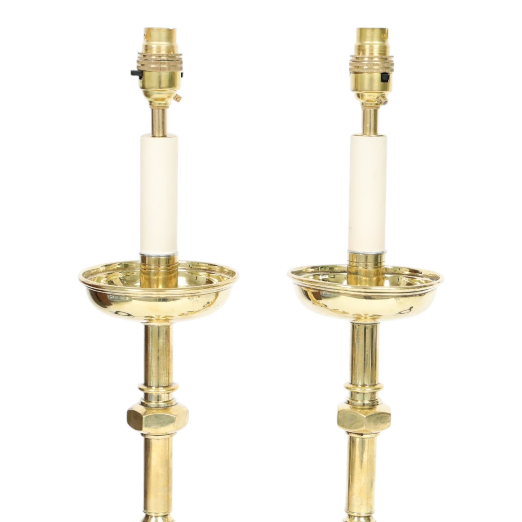 Pair of early 20th century brass alter candlesticks converted to table lamps, each with stepped and spreading circular base leading to a double hexagonal knopped column, and plain socket above inverted domed circular drip pan, the socket with light fitting, overall H49cm