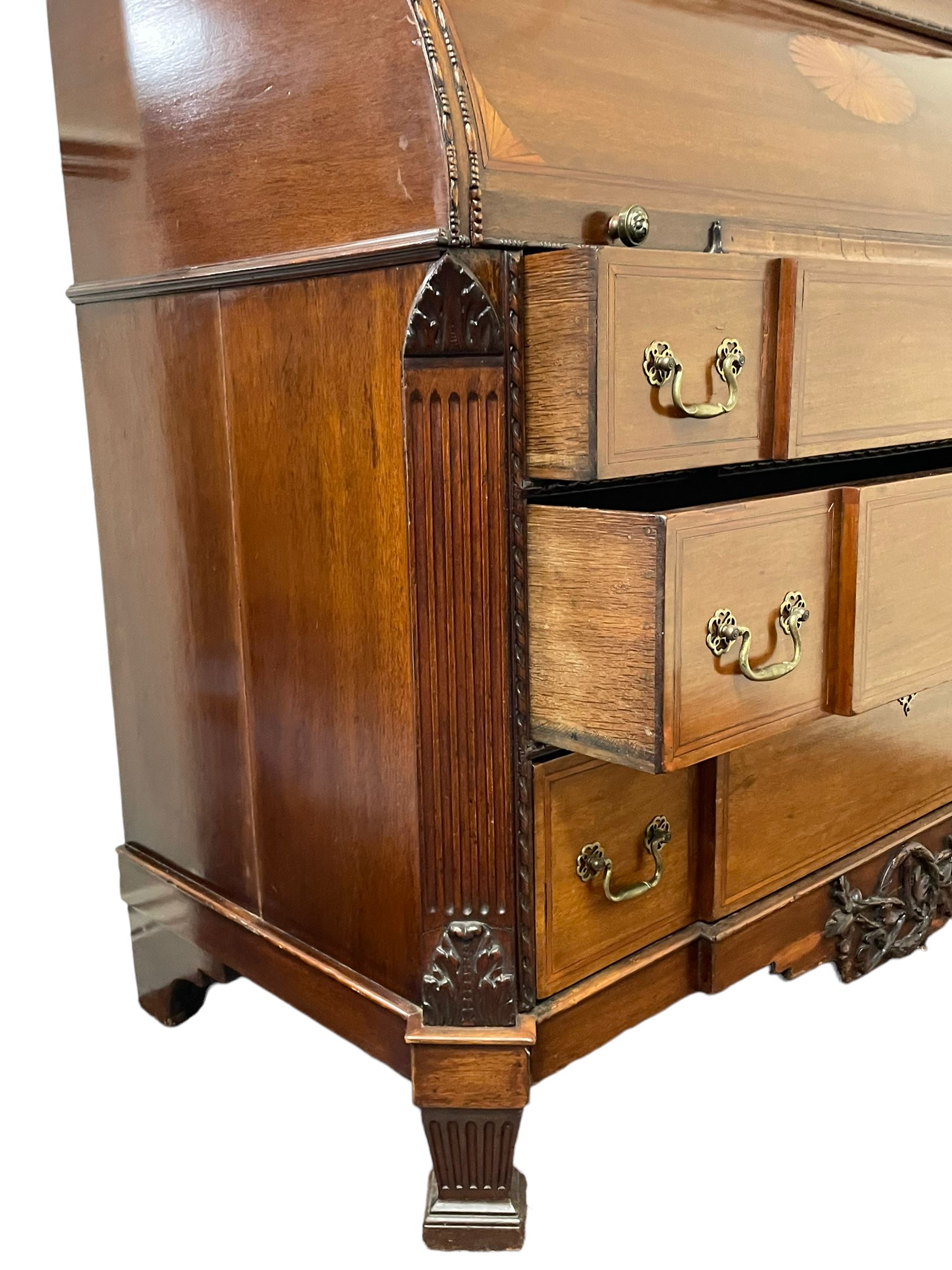 Large and impressive late 19th century Irish mahogany secretaire bookcase, moulded sloped arch dentil pediment decorated with central carved mask surrounded by linen festoons and trailing bellflowers, canted and fluted uprights with carved acanthus leaf upper and lower brackets, enclosed by two astragal glazed doors in the form of trophies with twist ribbon carved mouldings, inlaid with fanned spandrels and trailing leaf painted satinwood band, cylinder roll top inlaid with central fan motif and fanned spandrels, pull-out action revealing stepped fitted interior, central tambour roll cupboard flanked by two Composite pilasters and a combination of drawers and pigeon holes, fitted with three break-front drawers, matched canted and fluted corners with acanthus leaf carvings, on square tapering and fluted spade feet