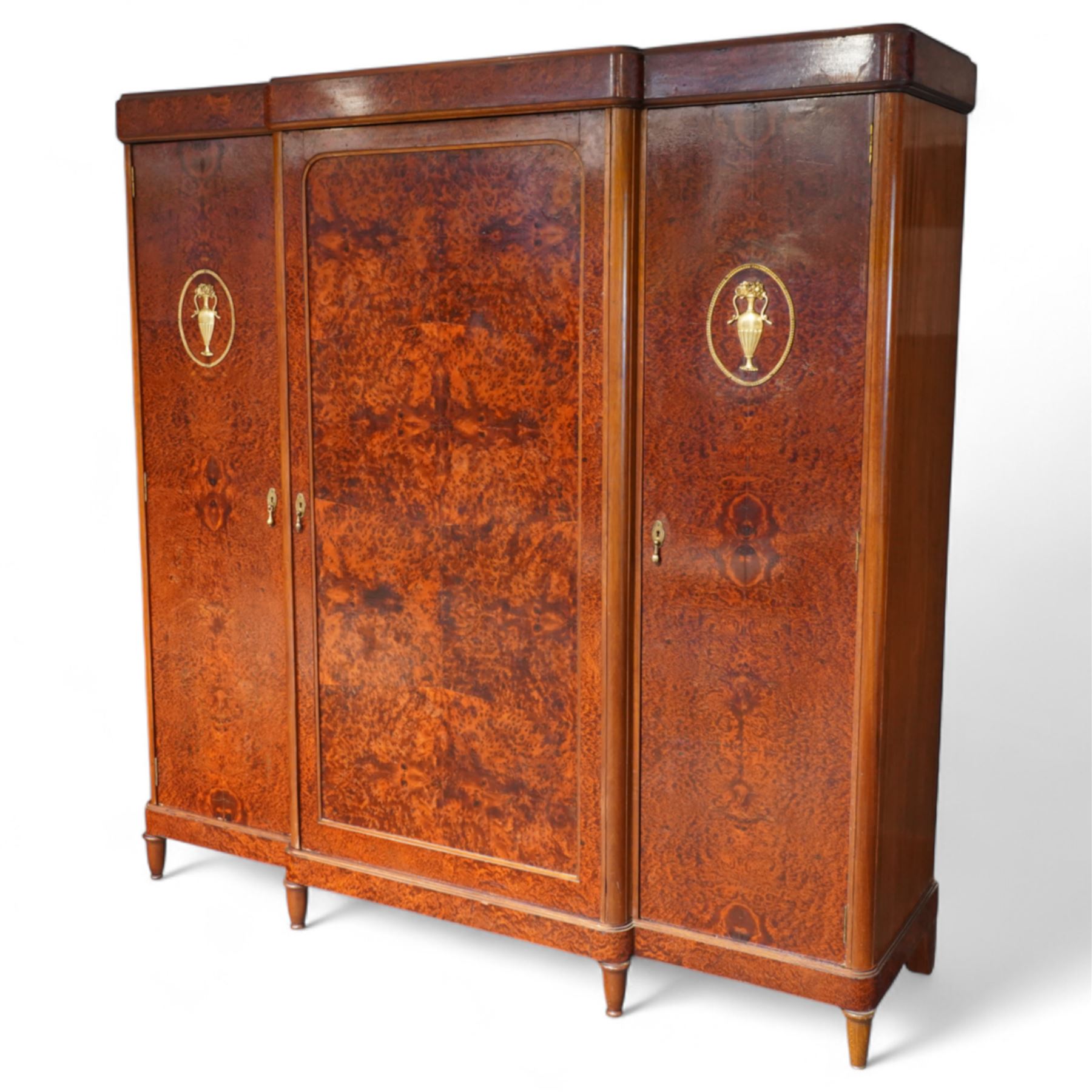 Early 20th century French burr maple breakfront armoire, enclosed by three panelled doors with bookmatched veneer and stringing, outer doors inlaid with gilt urn medallions, fitted interior with hanging spaces flanking a shelved compartment, raised on short tapered legs