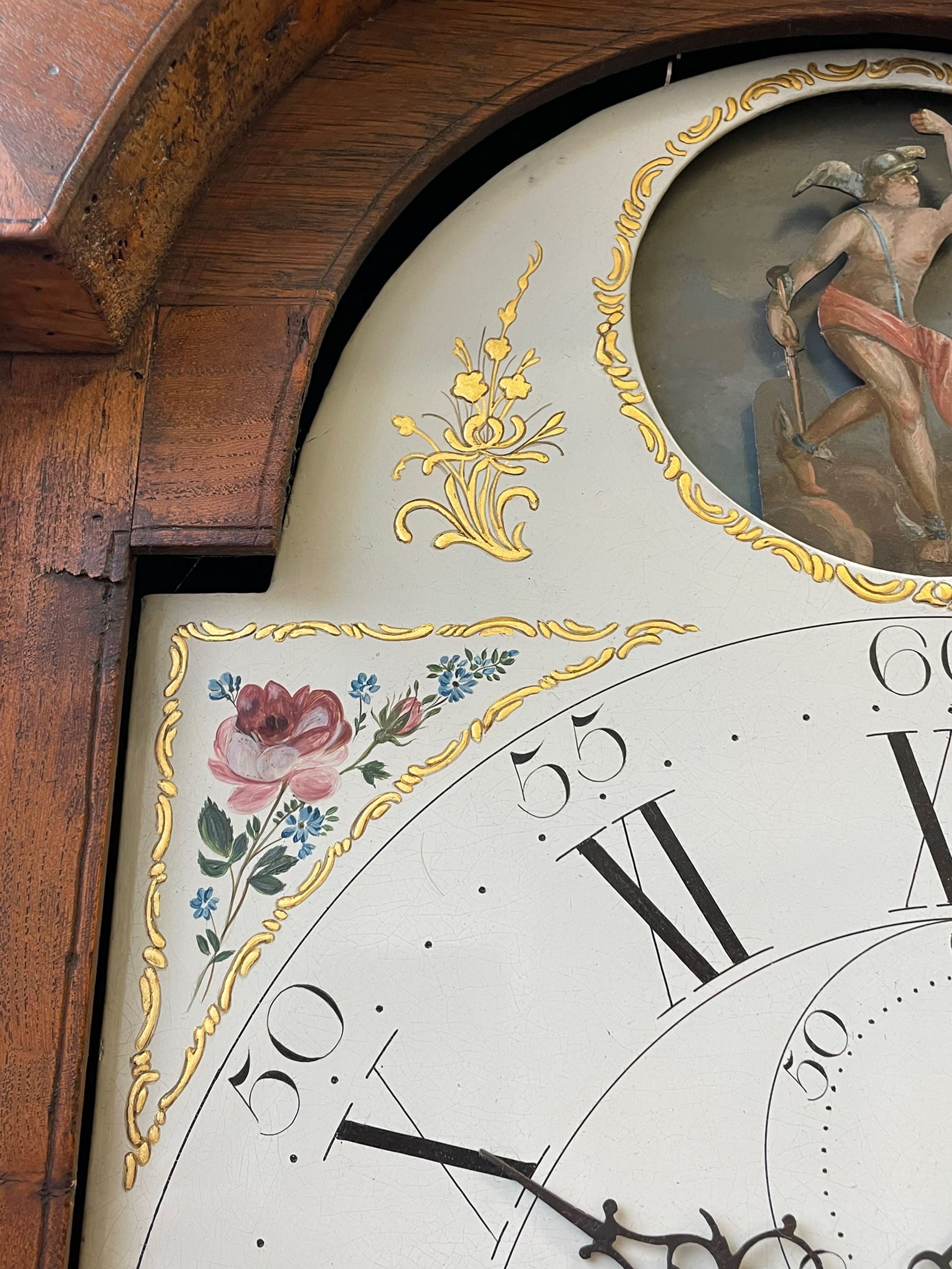 William Gostling of Diss  (Norfolk)-  Late 18th century 8-day oak longcase clock c1780-90, break arch top with scalloped cresting and three brass finials, glazed break arch hood door flanked by turned pilasters with brass capitals, trunk with a long trunk door and rounded top on a square plinth with an applied and shaped skirting, early painted Wilson of Birmingham dial with Roman numerals and five minute Arabic's, matching steel hands and second pointer, arched square ended calendar aperture with date disc behind, floral spandrels within gesso borders and a painted rocking automaton representing Mercury to the break arch, dial pinned via a false plate to a weight driven rack striking movement with a recoil anchor escapement, striking the hours on a cast bell. With pendulum weights and key. 
William Gostling (II) is recorded as working in Diss from 1774 to 1797, succeeded by widow Anne and son George . The dial of this clock is illustrated and discussed in detail in M.F. Tennants book 