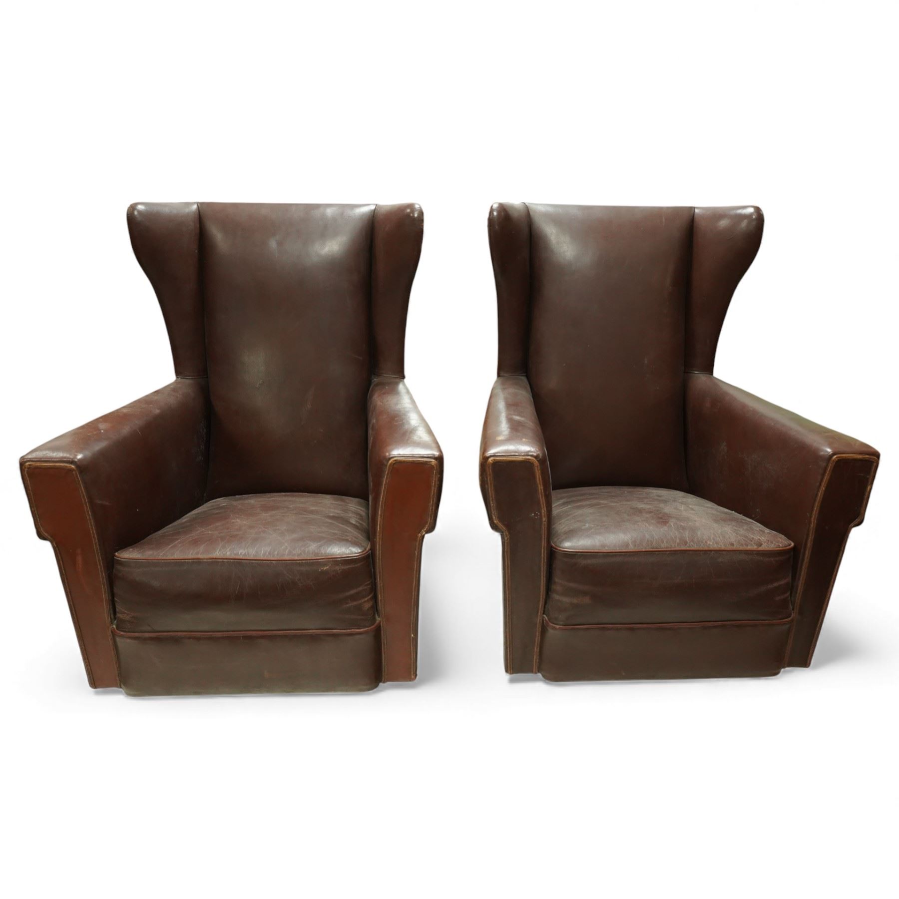 Early to mid 20th century pair of wingback armchairs, upholstered throughout in chocolate brown leather, the backs with deep wings, square padded arms with piped seams, loose seat cushions over solid bases