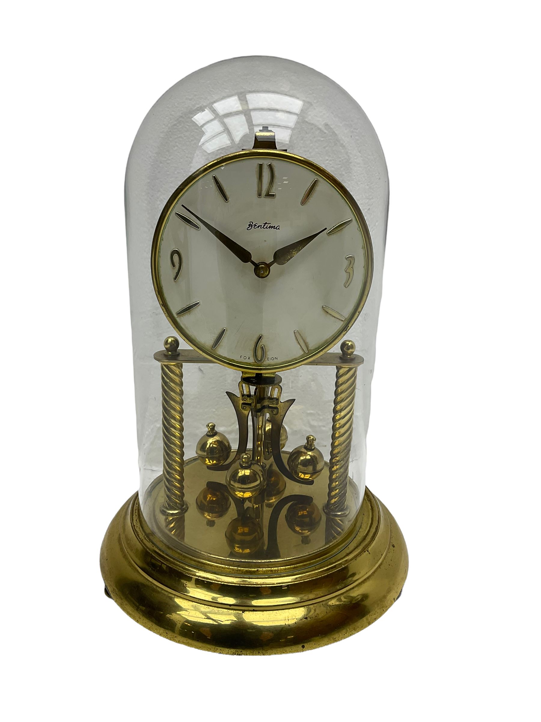 A mid-20th century “Bentima” 400-day anniversary torsion clock with a 7” circular brass base and unenclosed movement supported on two twisted columns, with a white dial gilt numerals and baton hands, four-ball rotary pendulum and glass dome. 

