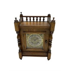 An early 20th century oak cased mantle clock in the arts and crafts style, case decorated with a rear balustrade, turned finials and flanking ring turned pilasters to the dial, square brass dial with cherub spandrels, silvered chapter ring, matted dial centre and gothic steel hands, with a German eight-day spring driven countwheel movement striking the hours and half hours on a coiled gong. With pendulum. 

