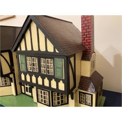 Large Tri-ang No.93 doll`s house in the 'Stockbroker's Range' c1935; two-storey double-fronted Tudor design, half timbered gables, two-storey bay windows, cream and mock shrubbery to front, opening metal framed windows with green shutters, red simulated tiled roof with chimney, integral garage with opening doors, front elevation has four hinged doors opening to reveal two bedrooms with fire places, living room, bathroom and kitchen with built in dresser and sink, entrance hall and staircase, side entrance with porch and seat and sun dial over on chimney stack; some wiring for electrical lighting; metal Tri-ang Toys disc verso; comprehensively furnished H66cm L117cm D43cm