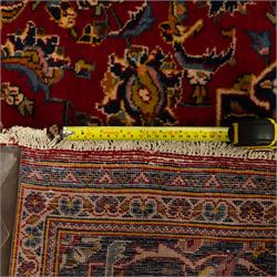 Central Persian Kashan hand-knotted carpet, crimson red field with a central shaped palmette medallion with pendants, filled with dense scrolling and floral motifs, enclosed by a navy blue border decorated with palmettes and meandering vine scrolls, flanked by multiple guard stripes, with cotton fringe ends 