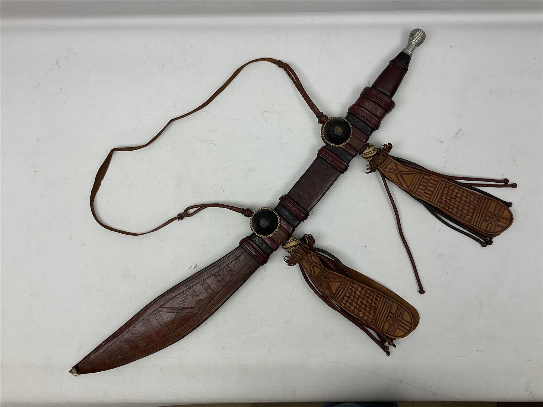 Mandingo sword with 63cm plain curving steel blade and leather covered grip with white metal ribbed spherical pommel; in decorative leather leaf scabbard with plaited tassels and shoulder strap L78cm overall