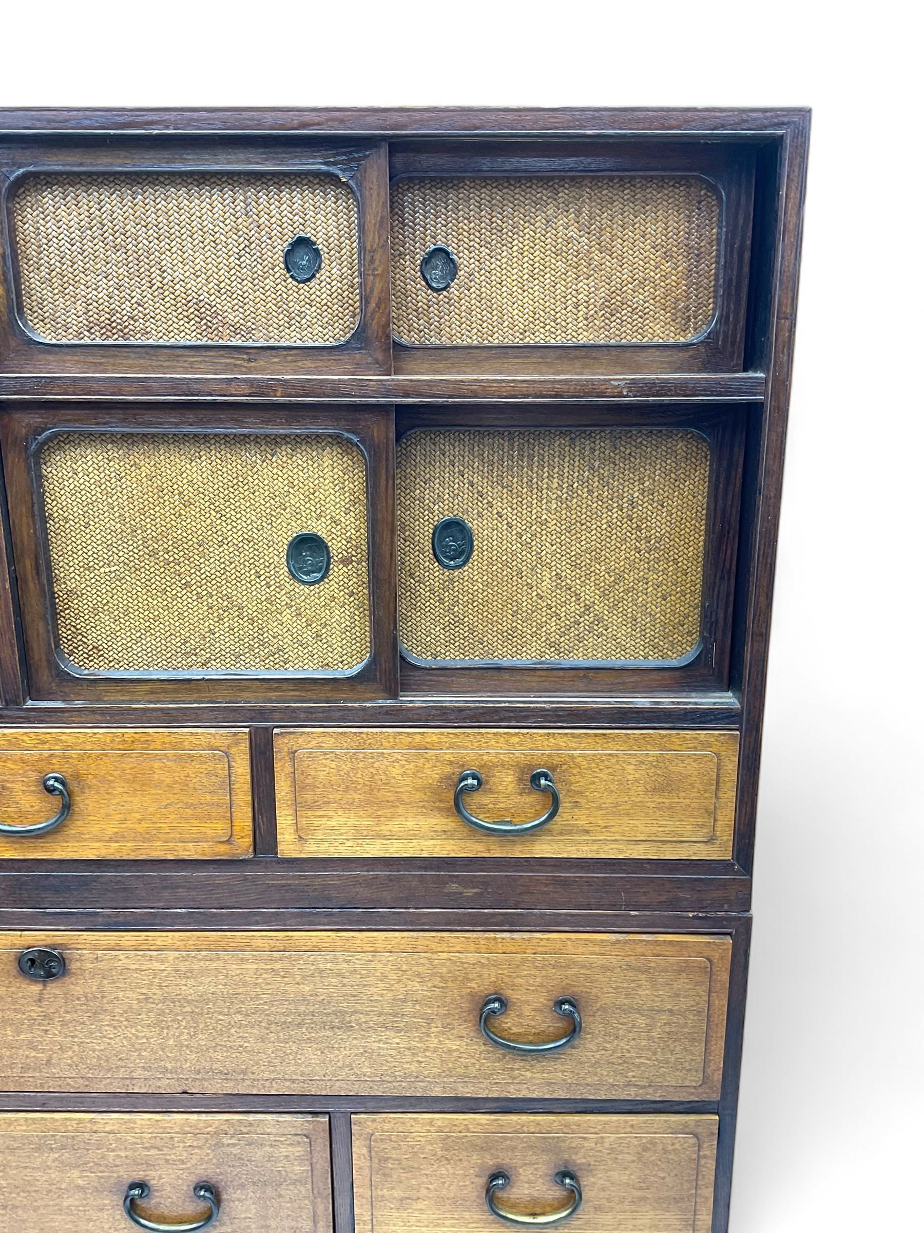 Late Meiji-period Japanese keyaki wood tansu chest, upper section with six sliding woven rattan doors over five drawers, lower section fitted with an asymmetrical arrangement of seven drawers and side cupboard with a hinged door enclosing three additional drawers, iron drop handles to sides