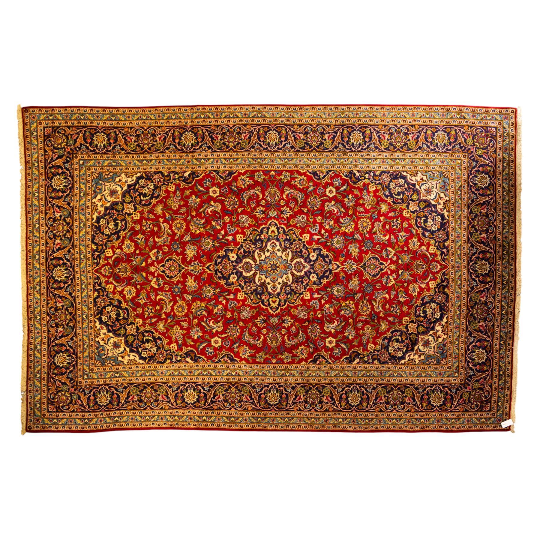 Central Persian Kashan hand-knotted carpet, central field in a ruby red ground with an indigo medallion with pendant motifs, enclosing scrolling arabesques, palmettes, and floral sprays in shades of indigo, turquoise, ivory, and gold, framed by indigo spandrels and a broad floral border between multiple guard stripes 