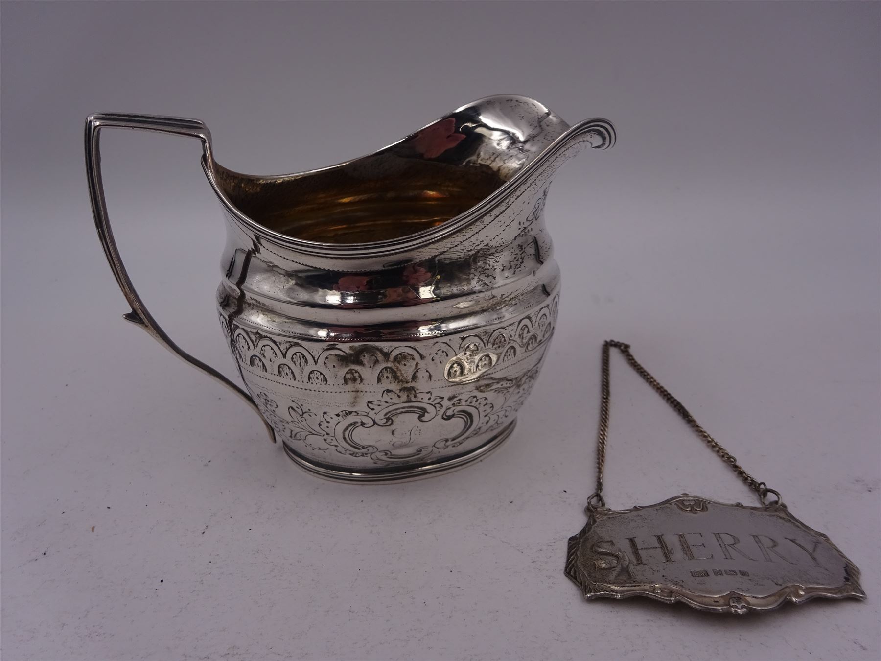 George III silver cream jug, of helmet form with angular handle, embossed with floral and scrolling decoration and engraved initial, hallmarked Edward Mayfield, London 1806, H9.3cm, together with a modern silver 'Sherry' decanter label, hallmarked Birmingham Mint, Birmingham 1986