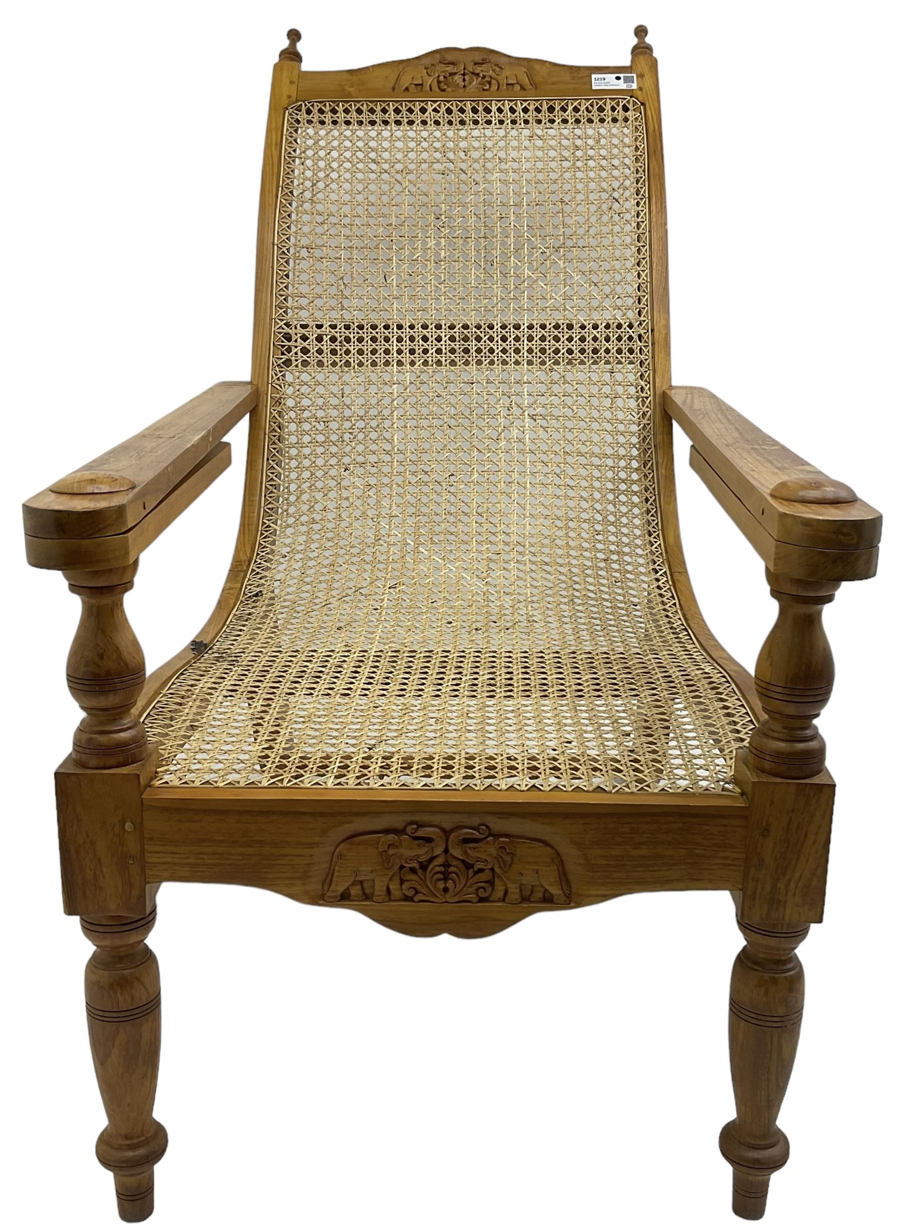 Late 20th century teak framed plantation chair, cresting rail and apron carved with two elephants with upraised trunks, flanked by finials, cane back and seat, fitted with hinged extending leg rests, on ring turned supports