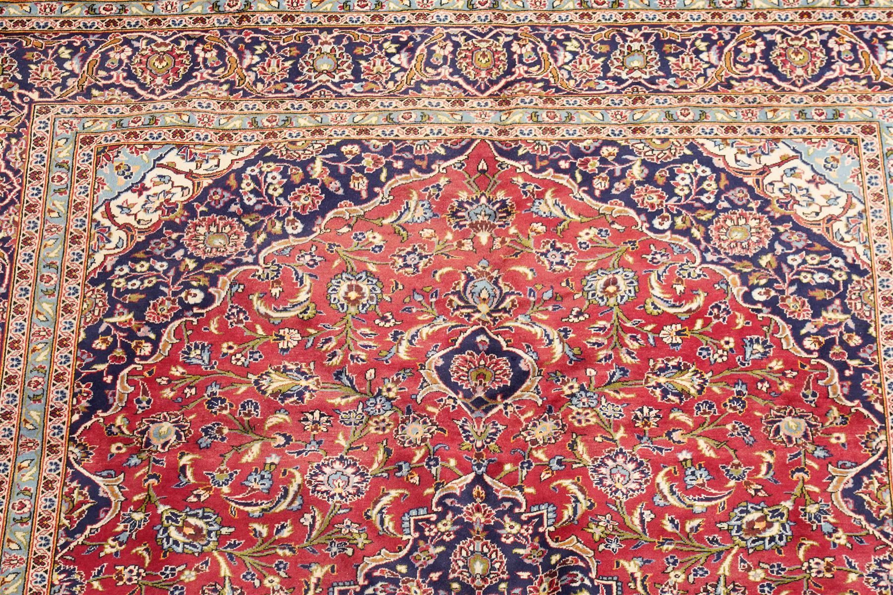 Persian Kashan hand-knotted carpet, madder red ground centred by shaped dark navy and ivory floral medallion with pendants, surrounded by scrolling vines, palmettes and flowerheads, dark navy spandrels, enclosed by dark navy floral border with multiple narrow floral guard stripes