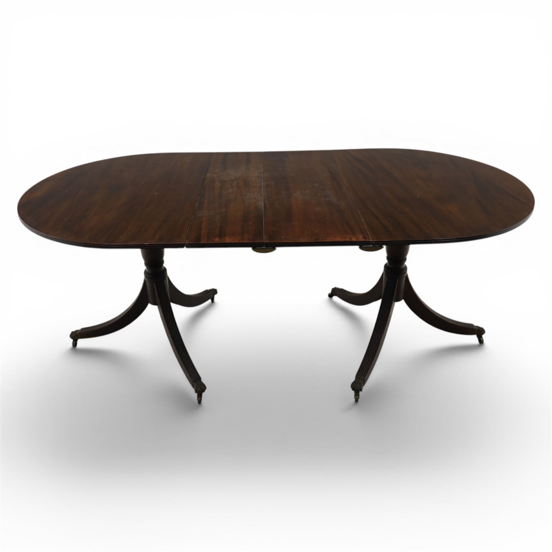 Set of eight George III style mahogany dining chairs, comprising two carvers and six singles, each with shaped top rail above pierced splay carved with feathers, with drop in stuffover seats upholstered in blue striped fabric, raised upon square tapering legs joined by stretchers, together with a mahogany twin pedestal dining table, the oval top with central leaf raised upon turned columns and splayed reeded legs terminating in lion paw feet and brass castors, 