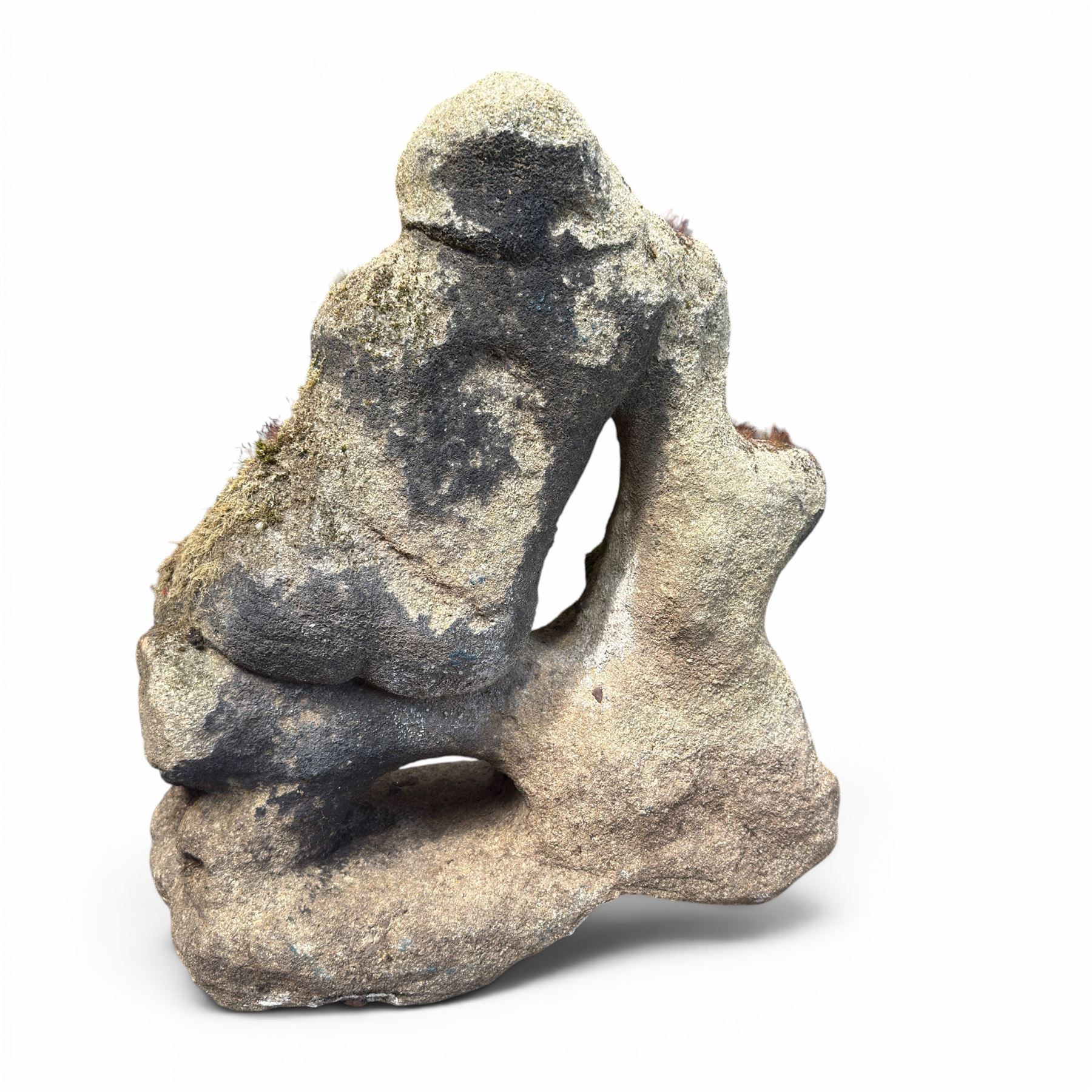 Carved composite stone garden figure, depicting a seated child with folded arms, weathered finish with moss and lichen growth, raised on an integral naturalistic base