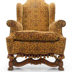 Pair of William & Mary design wingback armchairs, the arched high-back over rolled arms with a loose seat cushion and sprung seat, upholstered in old-gold ground foliate patterned fabric, raise on scroll feet united by a front serpentine stretcher