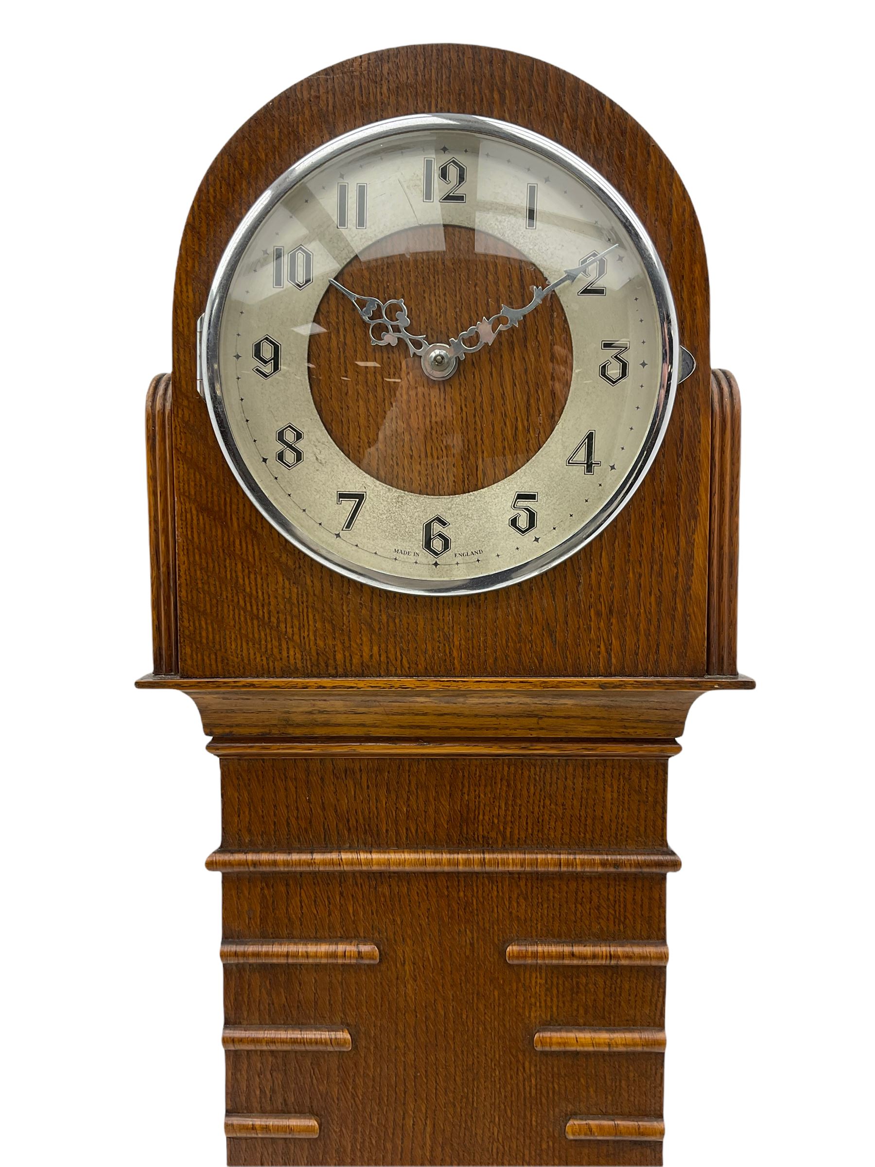 A mid-20th century English art-deco Grandmother clock with a mains powered electric movement chiming the hours and quarters on gong rods, in an oak case with an arched top, long trunk and short plinth on bracket feet, sliver effect chapter ring with Arabic numerals and silvered serpentine hands, with chime/silent facility.



