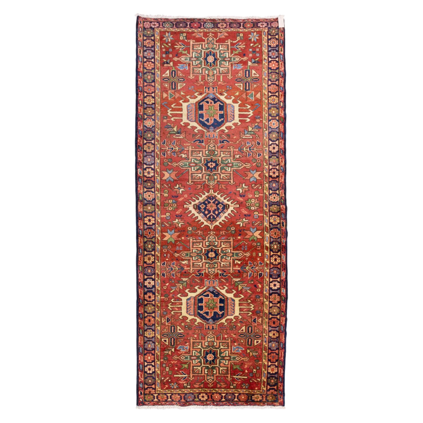 North West Persian Karajeh hand-knotted runner, madder red field, decorated with a vertical sequence of bold geometric medallions, interspersed with stylized floral and tribal motifs, framed by a dark ground border of angular palmettes and rosettes with multiple guard stripes, with cotton fringe ends 