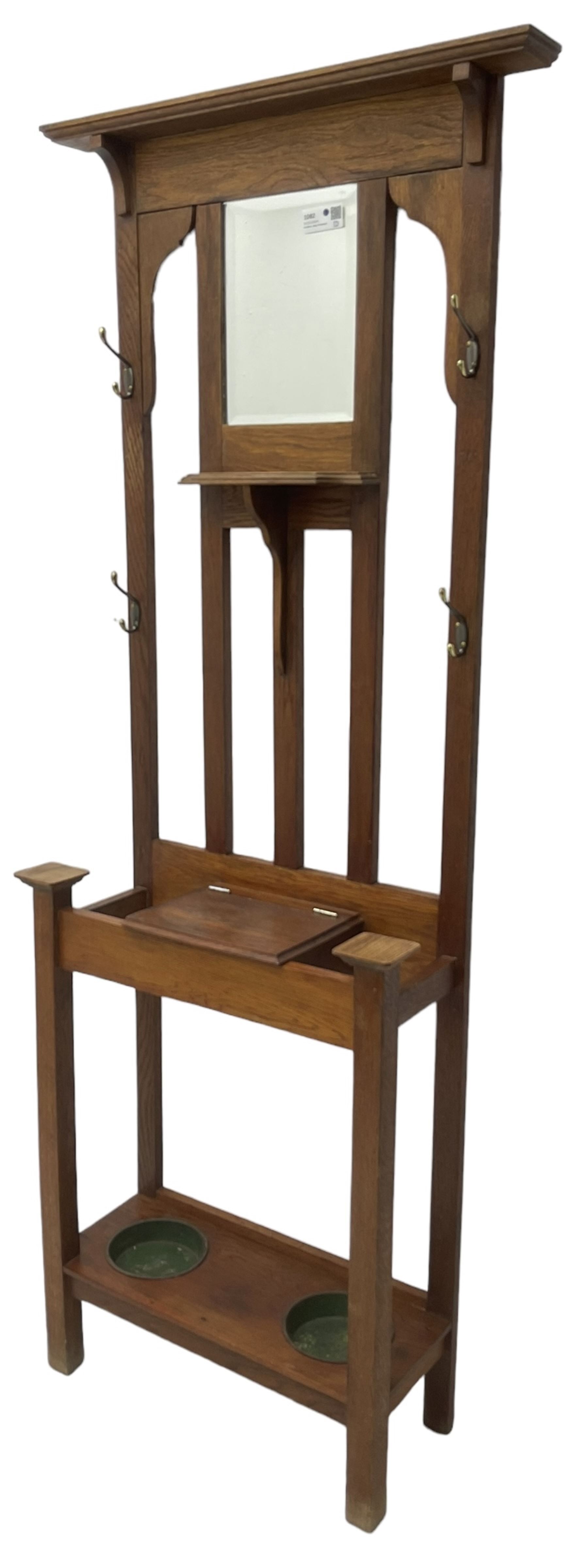 Edwardian oak hall stand, raised back with rectangular bevelled mirror plate surrounded by shelf and coat hooks, fitted with a glove box with hinged lid, over an undertier with two inset umbrella drip trays
