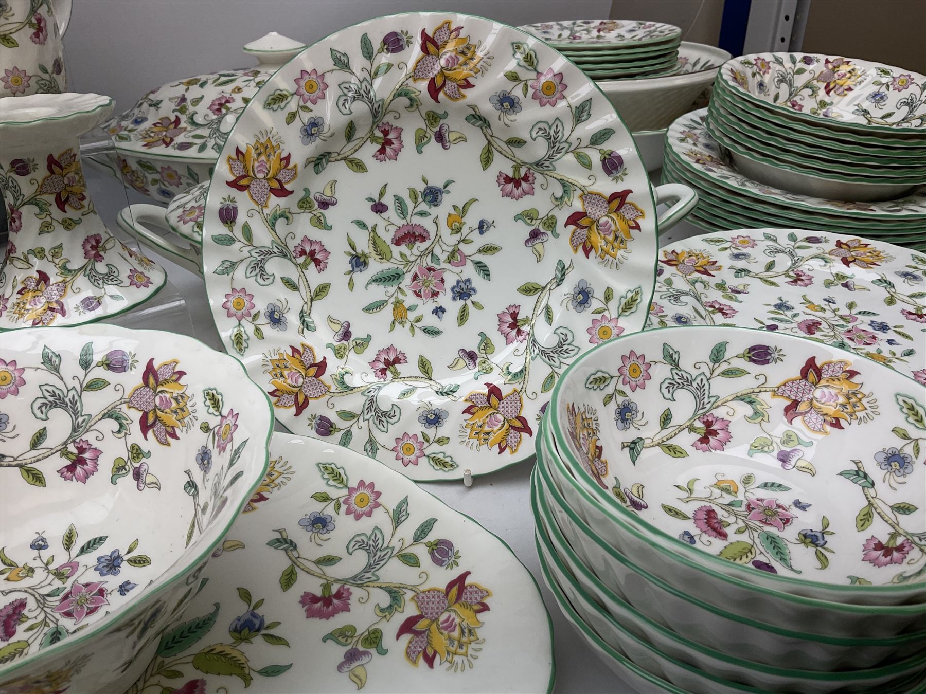 Minton Haddon Hall pattern tea and dinner wares, including basket dish, two covered tureens, six dinner plates, eight dessert plates, twelve tea plates, six soup bowls, six cereal bowls, teapot, coffee pot, milk jug, covered sucrier twelve tea cups and saucers, six mugs, footed bon bon dish, pair of candlestick holders, cake knife and slice etc (81)