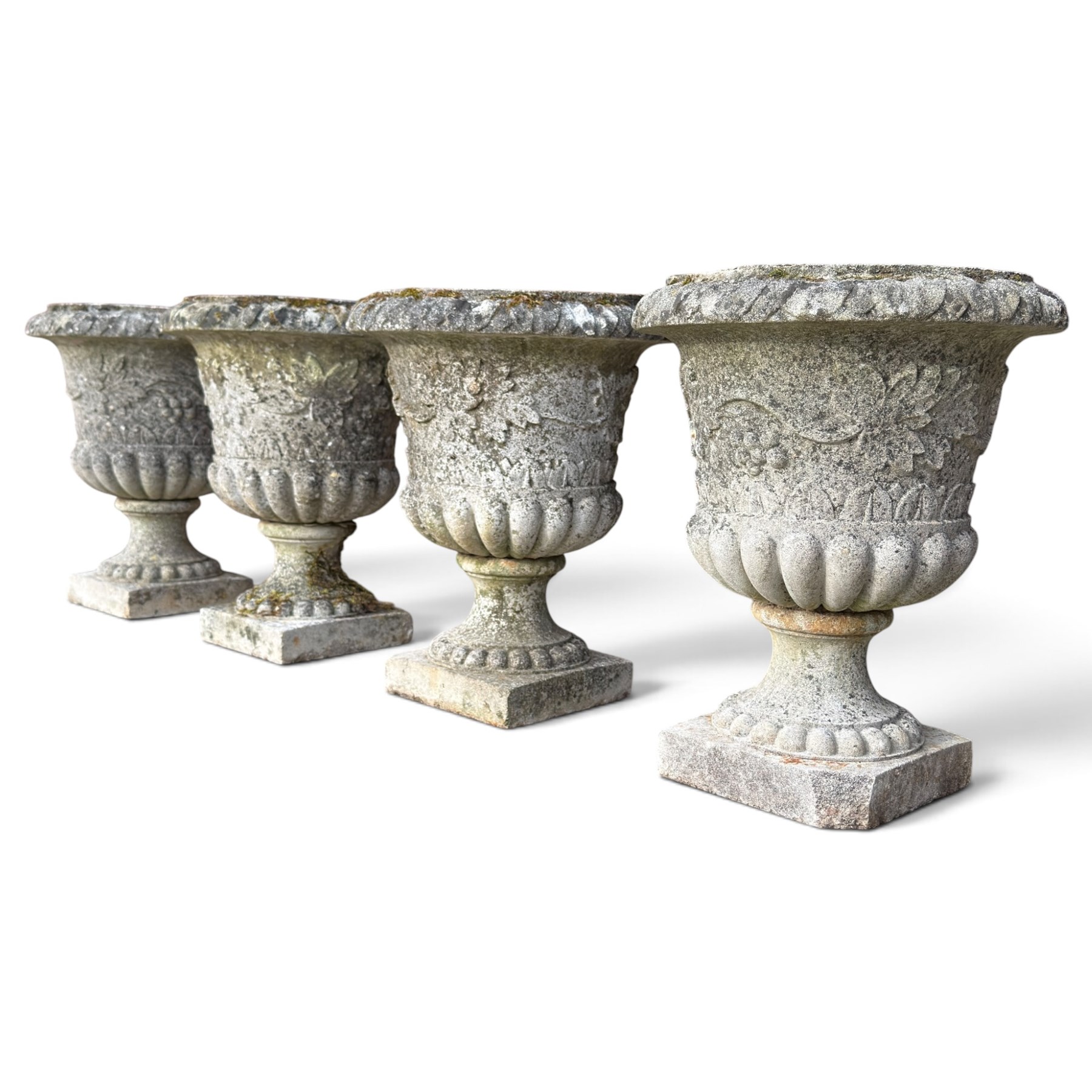 Set of four weathered composite stone urns of campana form, the everted leaf-moulded rolled rim over a body decorated with scrolling fruiting grapevines in relief, over a gadrooned underbelly and waisted socle, lobed collar on square base