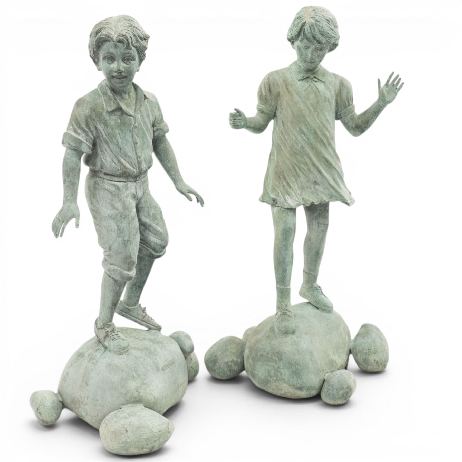 After Eneri Prosperi, bronze garden child statues, depicting children balancing on rock-form bases, the boy in shirt and shorts, the girl in dress with raised arms, surrounded by loose rounded pebble elements