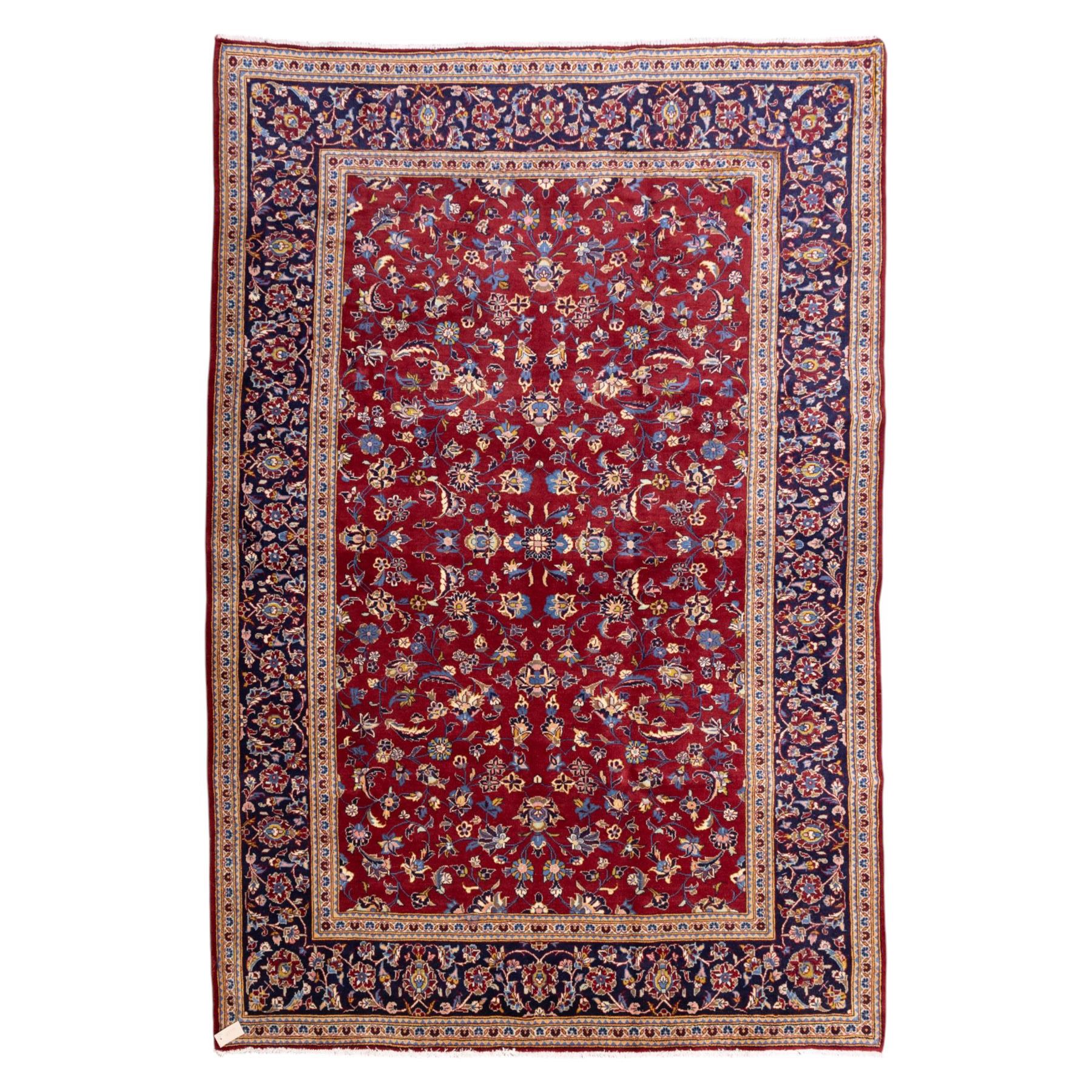 Central Persian Kashan hand-knotted rug, crimson field, decorated with an all-over floral and foliate design incorporating stylized palmettes, rosettes, and scrolling vine work, enclosed by a dark navy blue main border ornamented with flowering branches and palmettes flanked by multiple narrow guard borders with repeating floral motifs, with cotton fringe ends 