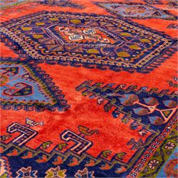West Persian Veis hand-knotted rug, coral red field decorated with a series of bold geometric lozenge medallions in shades of indigo and sky blue surrounded by angular tribal motifs and stylized animal and plant forms framed by a deep indigo border of geometric ornament and multiple guard stripes, cotton fringe ends 