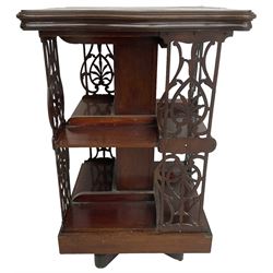 Edwardian mahogany revolving bookcase, the shaped moulded top inlaid with cusped panel and trailing bellflowers within a satinwood frame, foliate pierced upright splats and central square column supporting two tiers, on cruciform base with brass and ceramic castors 