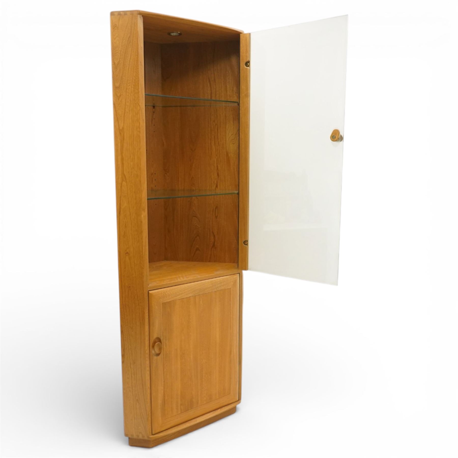Ercol - elm corner display cabinet, Model 3856. Glazed upper section with glass door enclosing two glass shelves and interior light, above panel cupboard door, with rounded uprights and circular turned handles, on plinth base