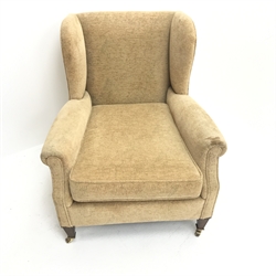  Edwardian low wingback armchair, upholstered in a beige fabric, square tapering supports brass castors, W75cm  