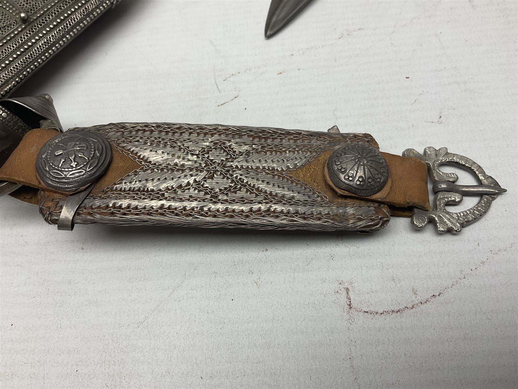 Omani Khanjar dagger, the 19cm curved blade with pronounced medial rib to either side, waisted hilt with dome shaped pommel and silver covered horn grip decorated with filigree bands, floral motifs and beadwork; in an intricately filigree decorated silver and leather scabbard with silver rings woven into the belt mount and fitted with original silver thread-work belt L34cm overall belt length 94cm