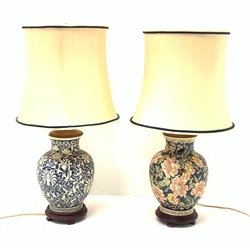 Two Oriental table lamps, each of bulbous form, the first example decorated with pink blossoming flowers upon a dark blue ground, above a fret pattern band, upon a wooden circular stepped base, the second example with white foliate decoration upon a dark blue ground, also with fret pattern band, and upon a wooden circular stepped base, each with fabric shade, including shade each approximately 74cm. 
