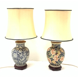 Two Oriental table lamps, each of bulbous form, the first example decorated with pink blossoming flowers upon a dark blue ground, above a fret pattern band, upon a wooden circular stepped base, the second example with white foliate decoration upon a dark blue ground, also with fret pattern band, and upon a wooden circular stepped base, each with fabric shade, including shade each approximately 74cm. 

