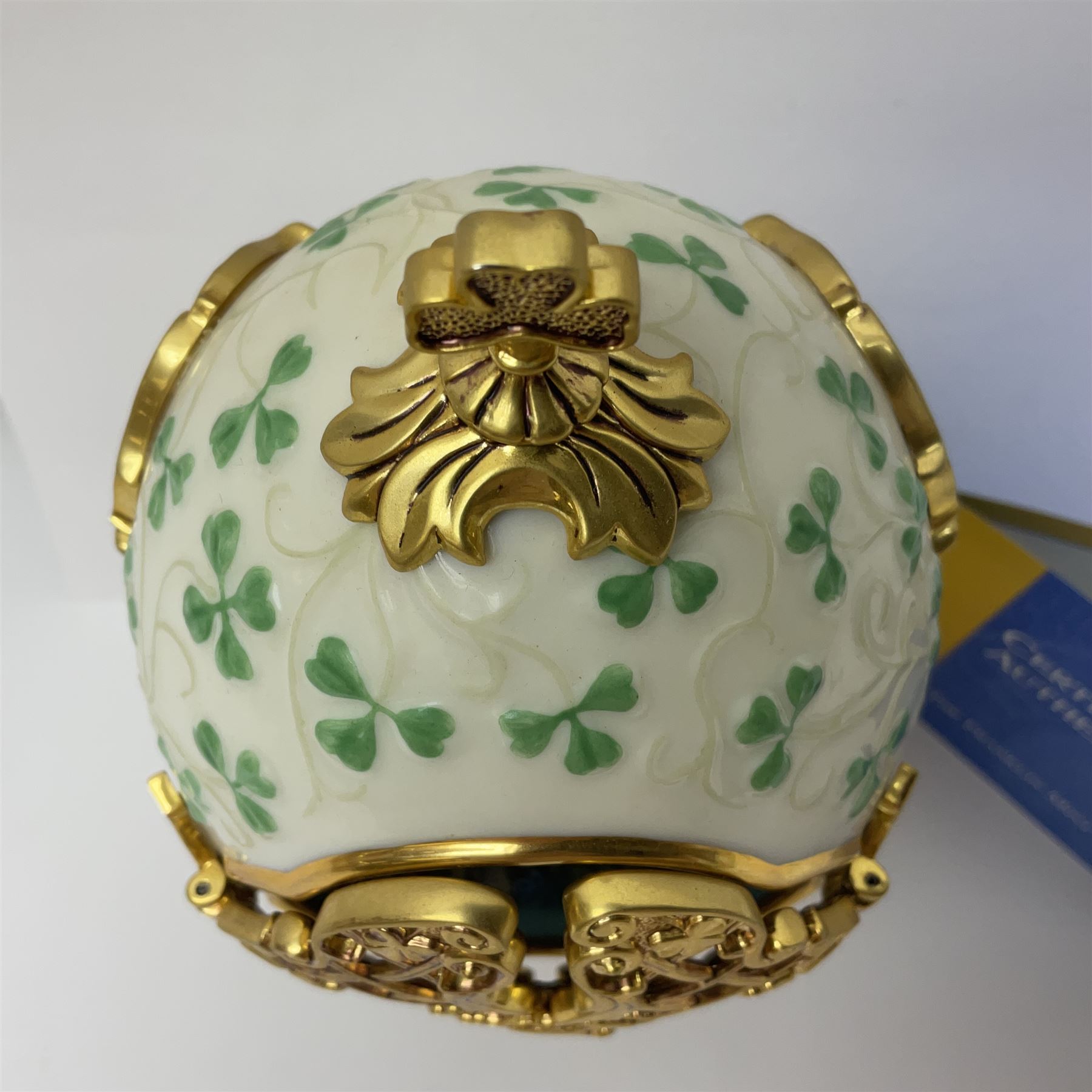 Franklin Mint The Emerald Isle Collector Egg, the egg decorated with embossed shamrocks upon a cream ground, with a gilded shamrock finial, accents and openwork door, opening to reveal a miniature landscape scene, upon three gilded feet set with green paste stones, with certificate of authenticity H23cm, 