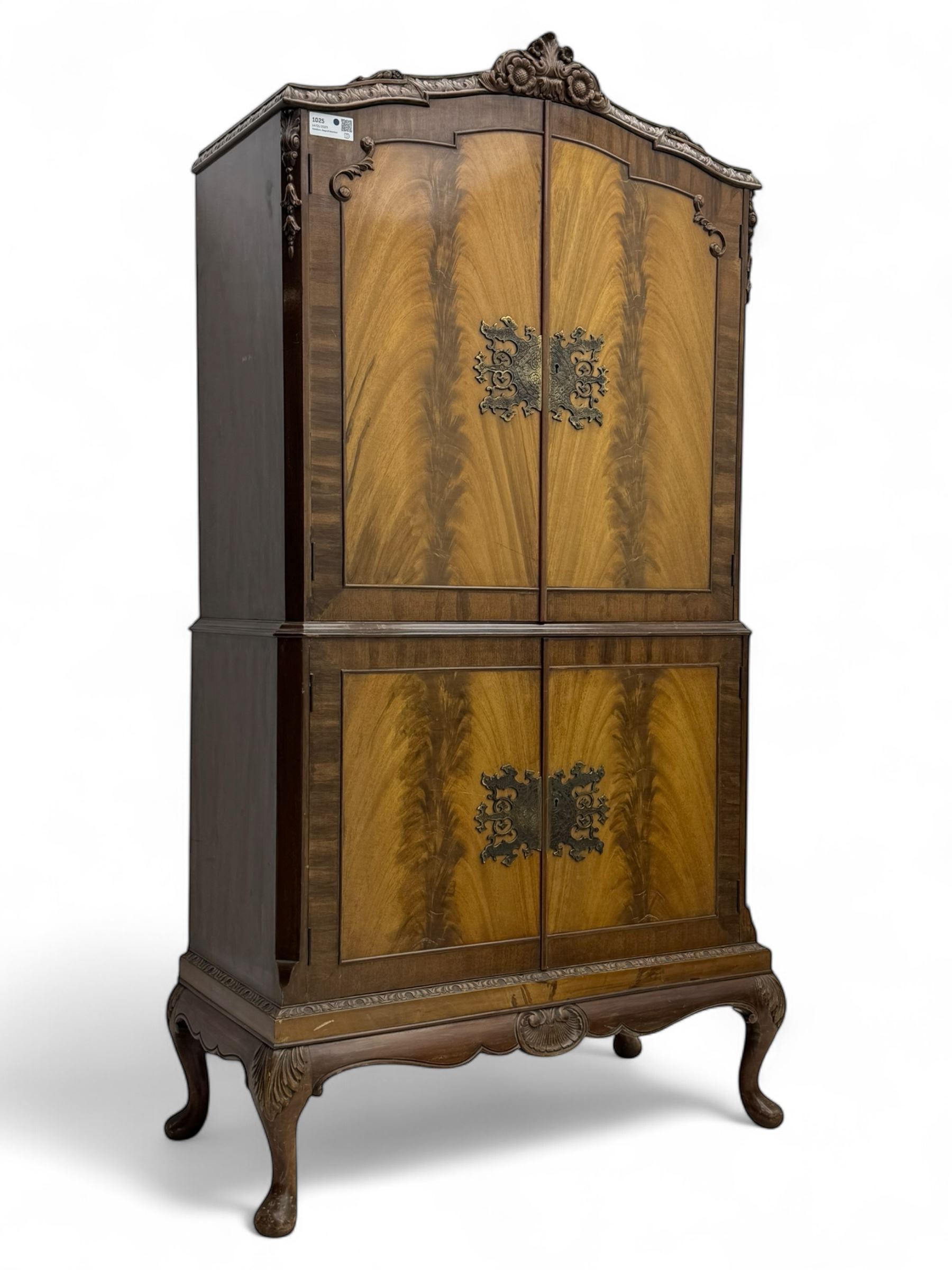 Early 20th century walnut veneered cocktail cabinet, shaped and carved foliate pediment above a pair of arched panelled doors with figured veneers and ornate cast escutcheons, interior is mirrored on the base and back, slide out mirrored shelf, flanked by foliate carved uprights, over a conforming lower cabinet, all raised on shaped apron and scroll carved cabriole feet