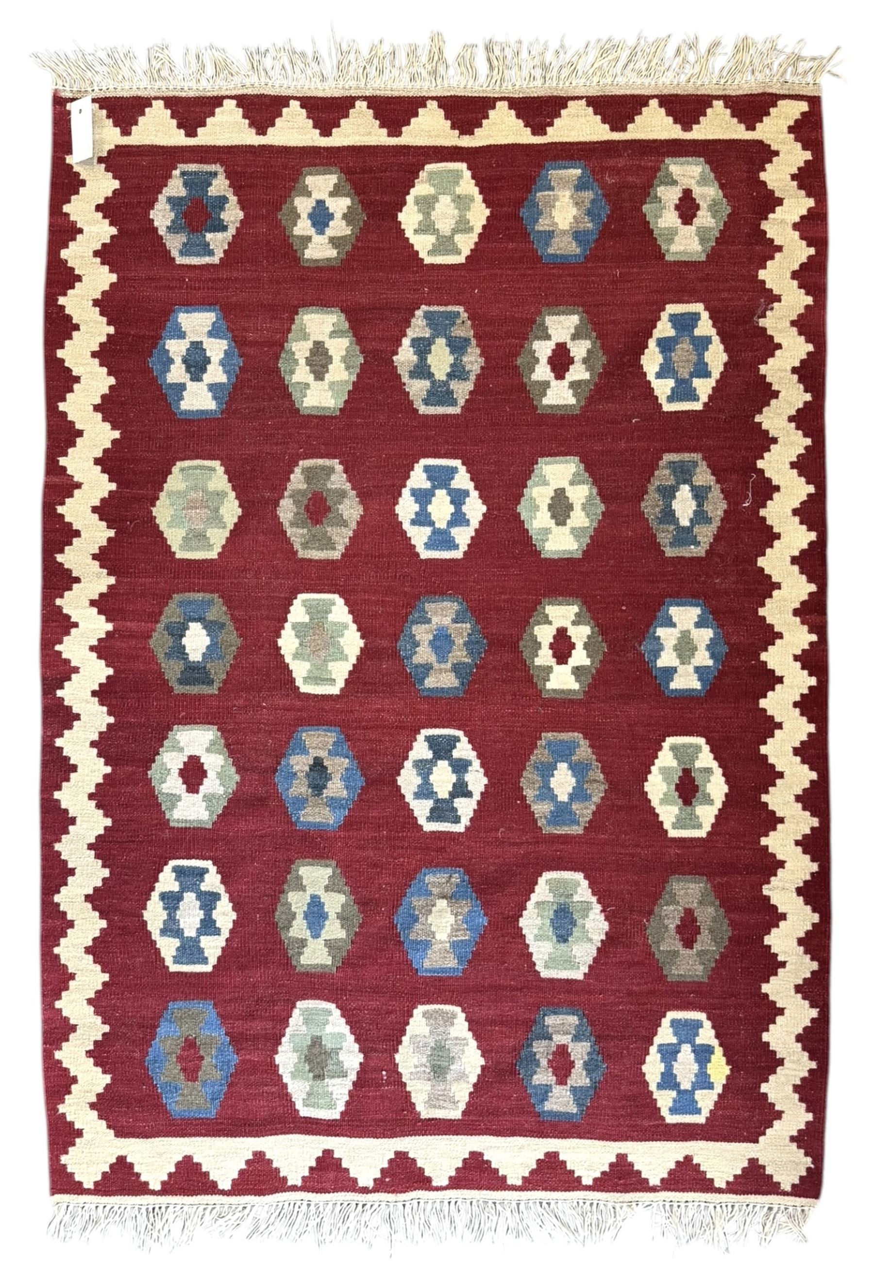 Handwoven wool Kilim rug of rectangular form with cream fringe, the red ground decorated with repeated multicoloured hexagonal medallions within a cream stepped border