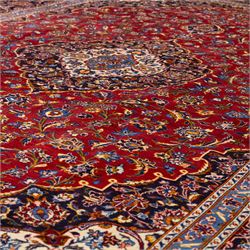 Central Persian Kashan hand-knotted carpet, rich crimson red field with a central cusped medallion with finely drawn floral sprays surrounded by dense scrolling vine work and palmettes, framed by a broad indigo ground border of flowering arabesques flanked by multiple guard stripes, in a palette of red, blue, ivory, sage green, and soft ochre tones, with cotton fringe ends    