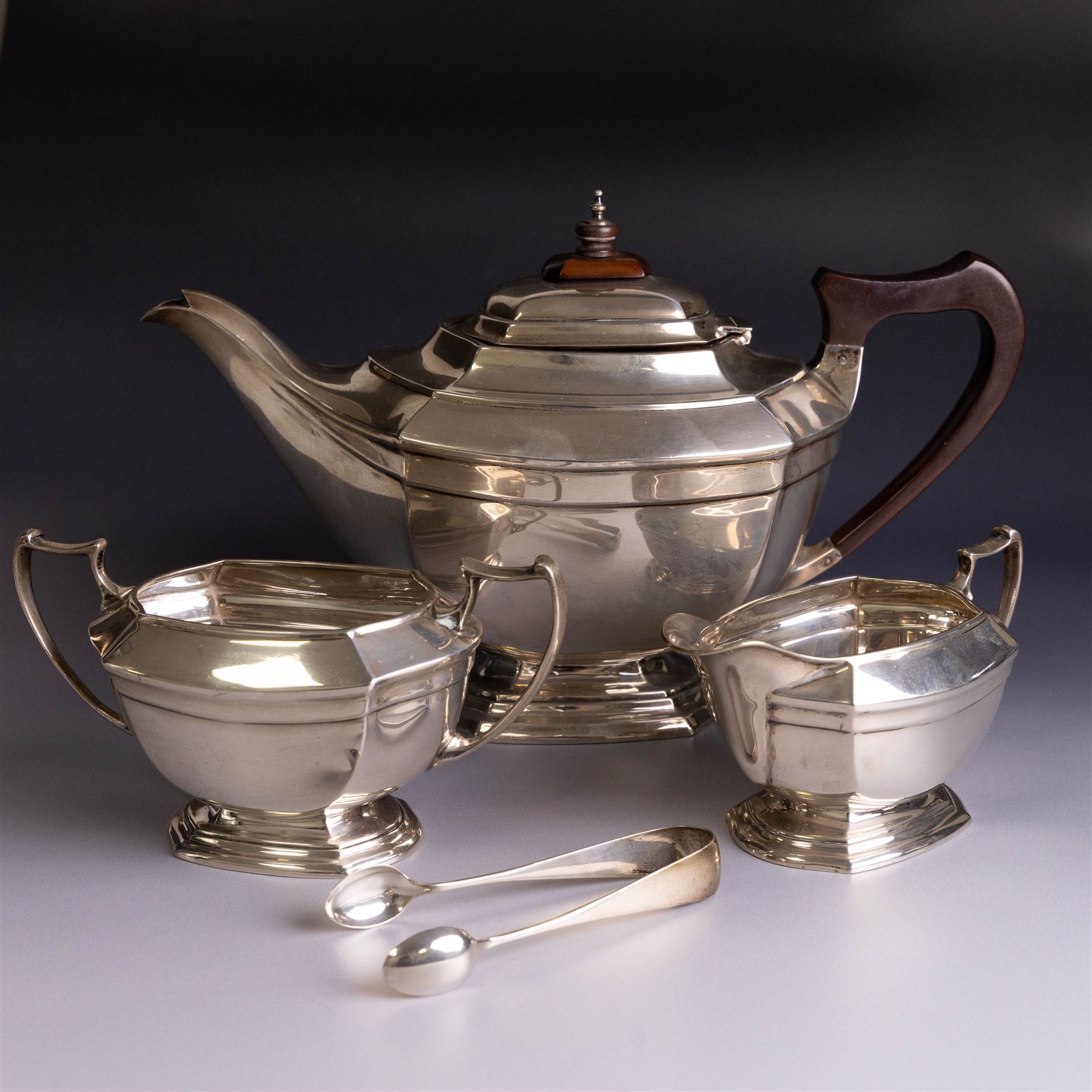 Art Deco three-piece silver tea service, comprising teapot with removable tea strainer, open sucrier and milk jug, all of hexagonal form, the teapot with wooden handle and finial, all hallmarked James Deakin & Sons, Sheffield 1925, together with a matched pair of silver sugar tongs, hallmarked