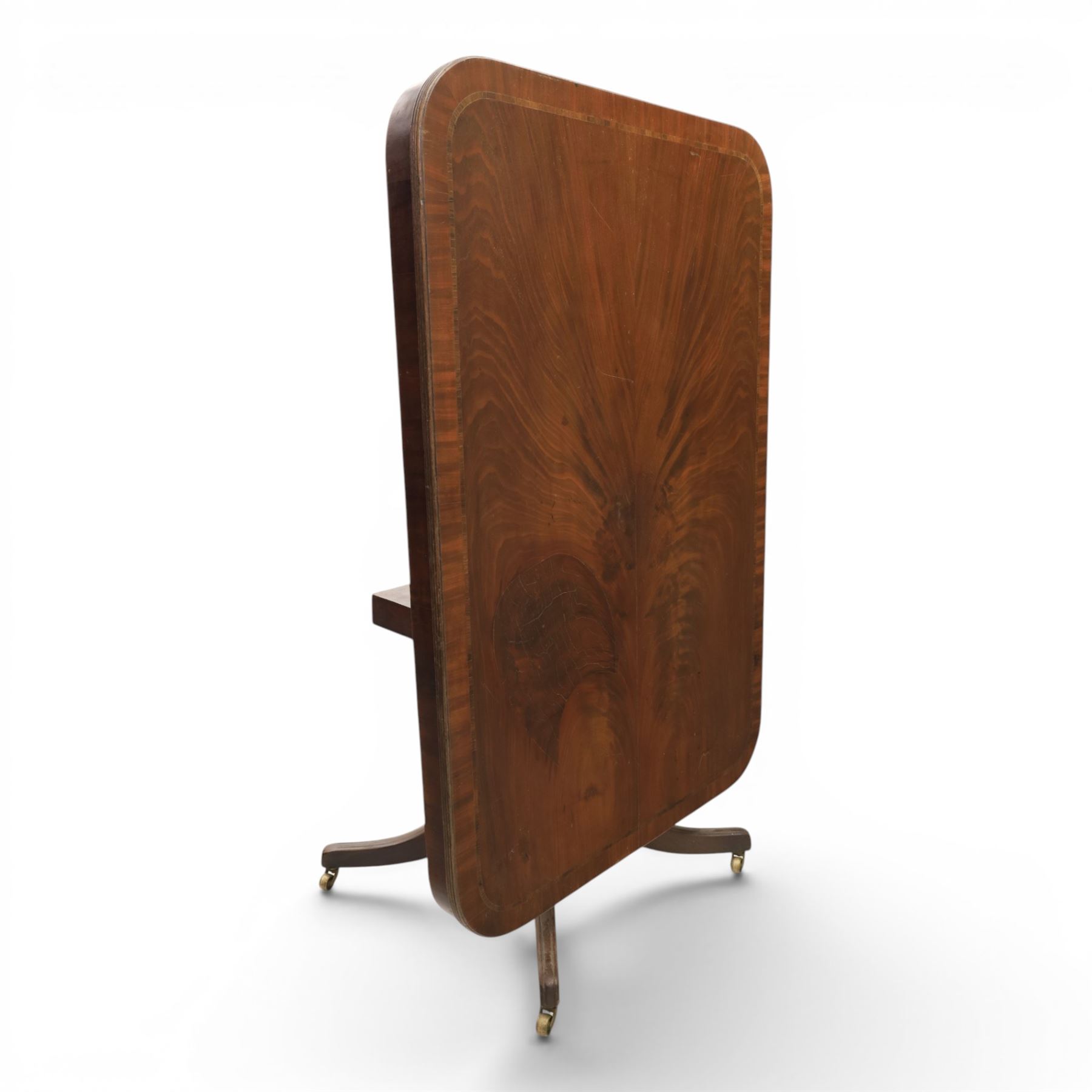 Regency mahogany tilt-top dining table, crossbanded rounded rectangular top, raised on a splayed base, terminating in brass castors