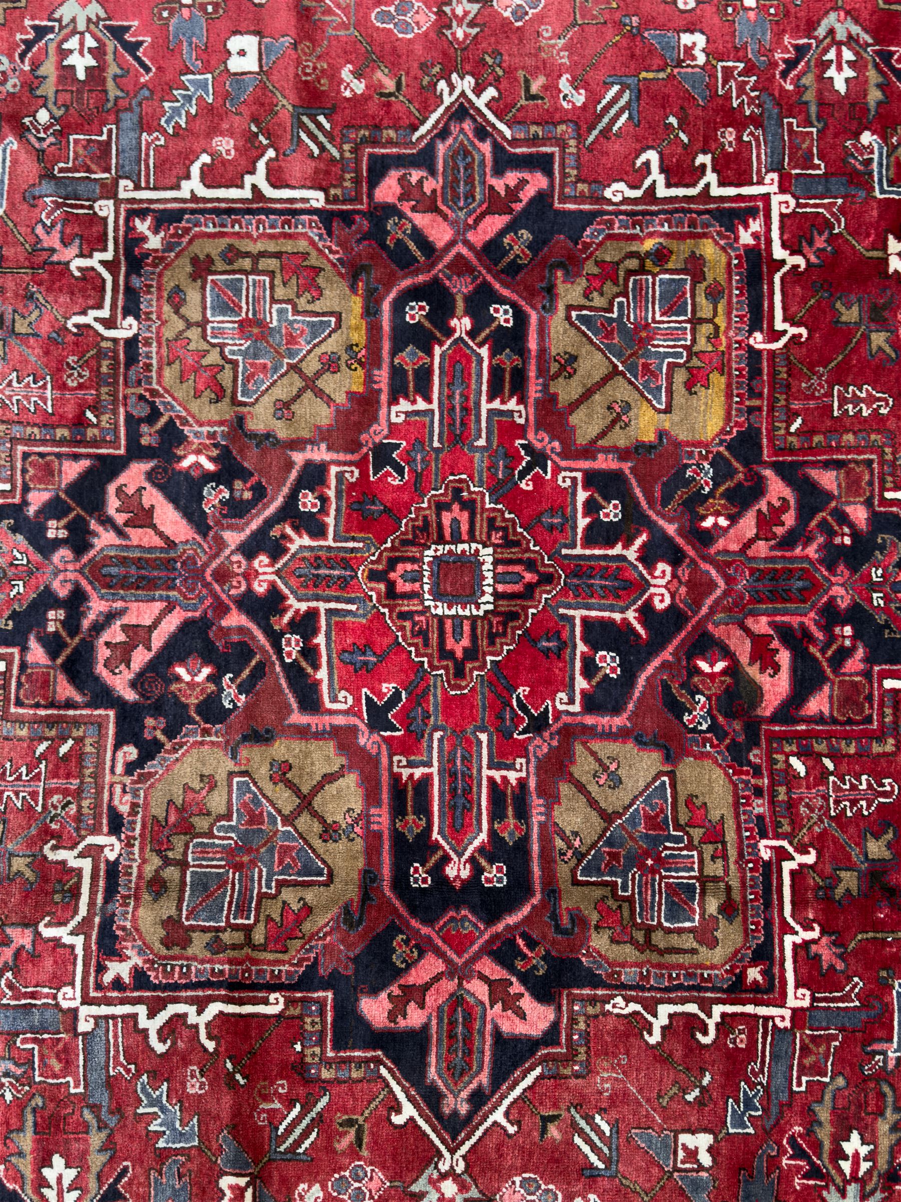Northwestern Persian Heriz hand-knotted carpet, madder red field with a bold stepped medallion in indigo, ivory, and ivory, surrounded by angular floral motifs and stylized leaves, and the spandrels set within a dark border of rosettes and angular vine motifs, 335cmx252cm