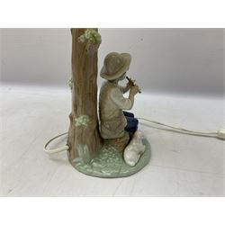 Nao table lamp modelled as a boy playing the flute upon logs with two recumbent sheep at his feet, the stem modelled as a tree trunk, with fabric shade, with printed mark beneath, H46cm