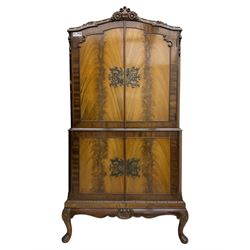 Early 20th century walnut veneered cocktail cabinet, shaped and carved foliate pediment above a pair of arched panelled doors with figured veneers and ornate cast escutcheons, interior is mirrored on the base and back, slide out mirrored shelf, flanked by foliate carved uprights, over a conforming lower cabinet, all raised on shaped apron and scroll carved cabriole feet
