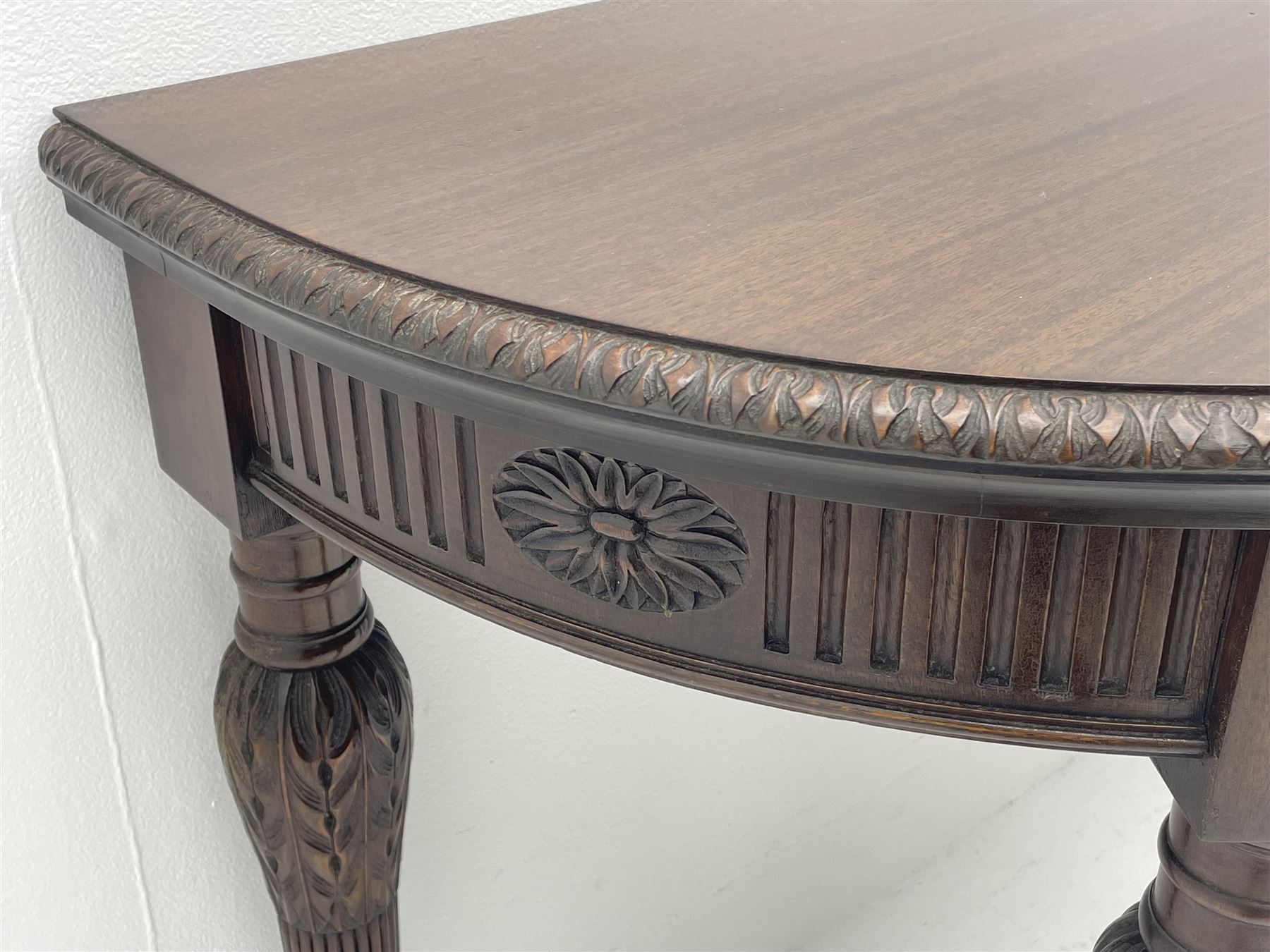 Pair early 20th century Hepplewhite style mahogany D-end console tables, the curved top with foliate carved edge, fluted frieze rails carved with flower heads, turned and reeded acanthus supports with recessed castors